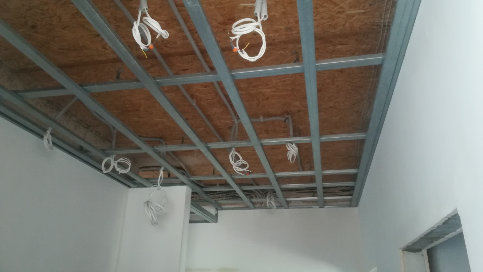 Stelaż metalowy podwieszony do sufitu z widocznymi przewodami elektrycznymi przed montażem płyt, widok z dołu w trakcie prac wykończeniowych.