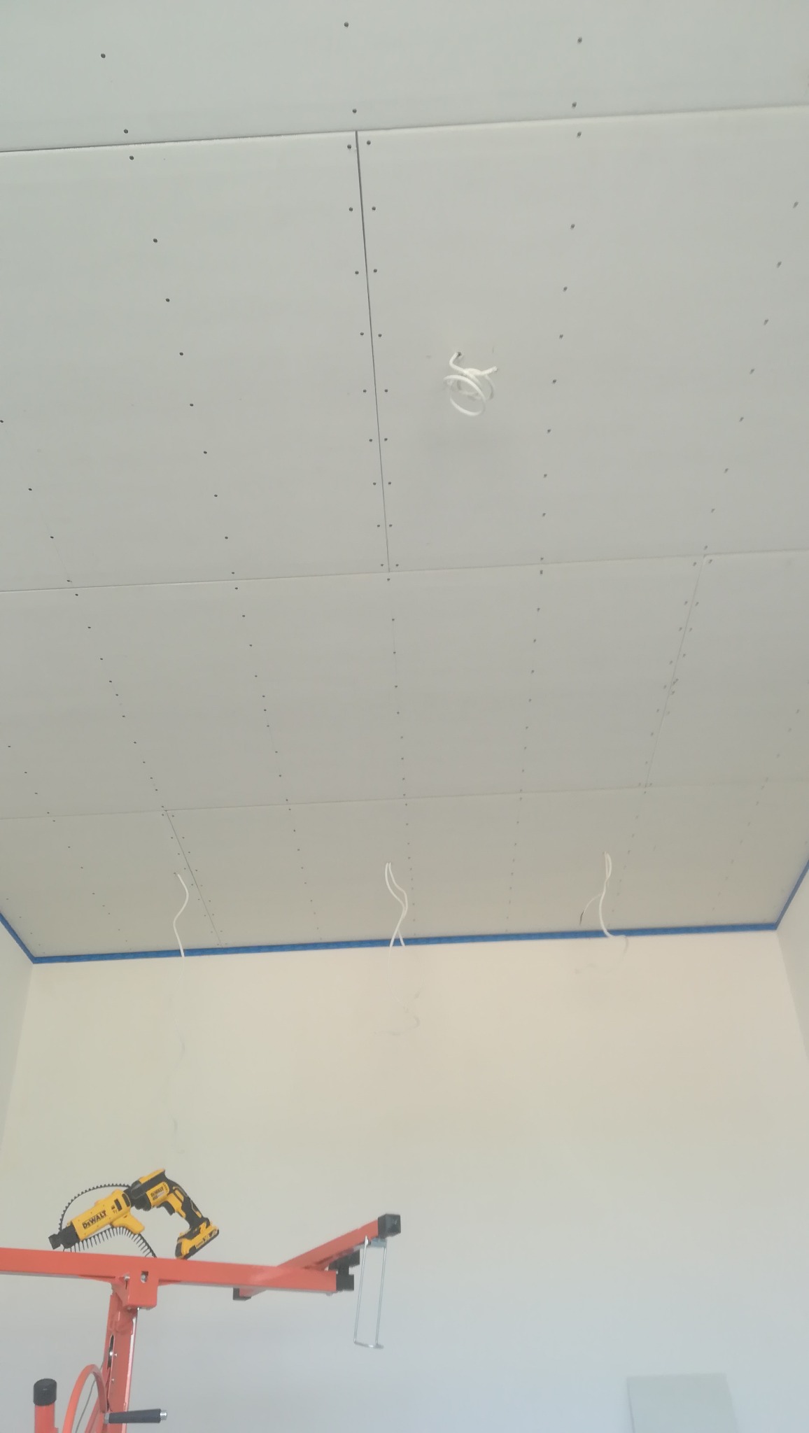 Montaż sufitu z płyt gipsowo-kartonowych, widoczna wkrętarka DeWalt na podnośniku, kable oświetleniowe wystające z sufitu i zabezpieczenie taśmą malarską przy ścianach.