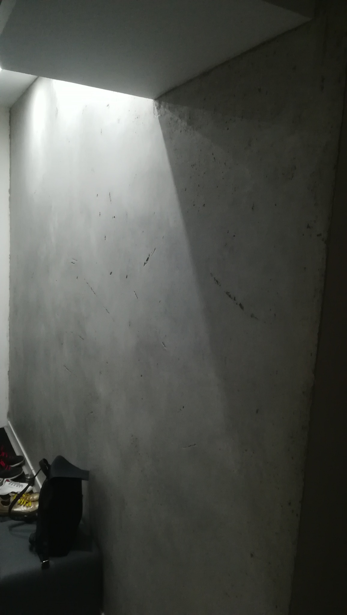 Ściana po nałożeniu szarej warstwy wykończeniowej, widoczne niedoskonałości i nierówności, oświetlenie górne podkreśla fakturę.