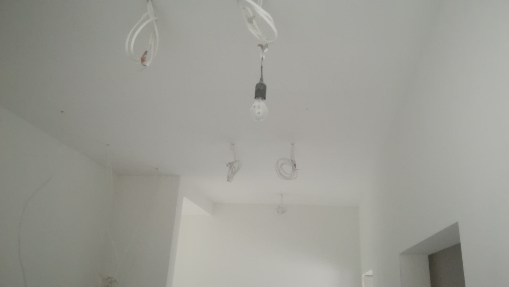 Wnętrze w trakcie remontu z widocznymi kablami elektrycznymi wystającymi z sufitu i pojedynczą żarówką, ściany świeżo pomalowane na biało.