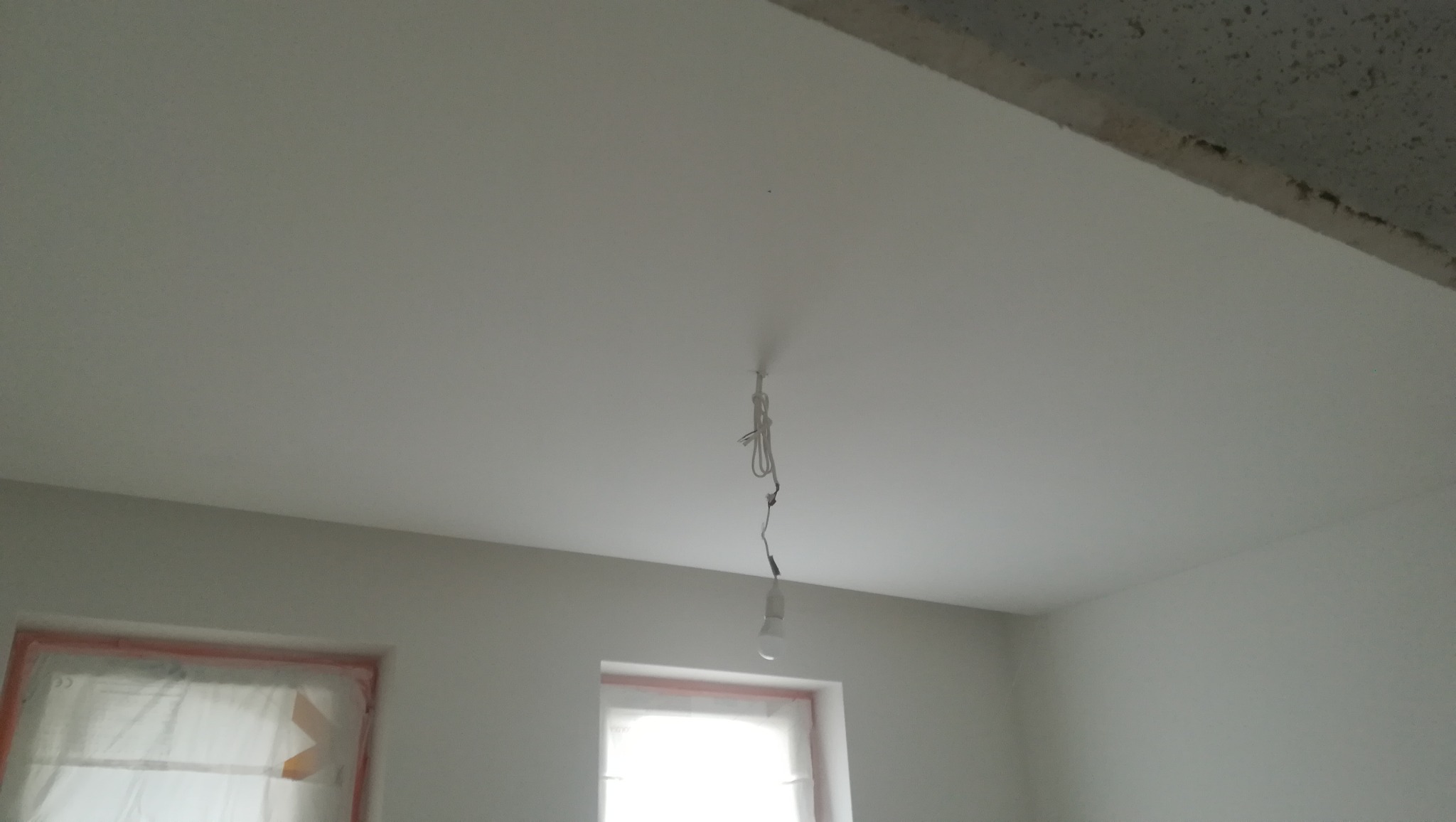 Świeżo pomalowane wnętrze z zabezpieczonymi folią oknami, sufit z wiszącą żarówką na kablu.