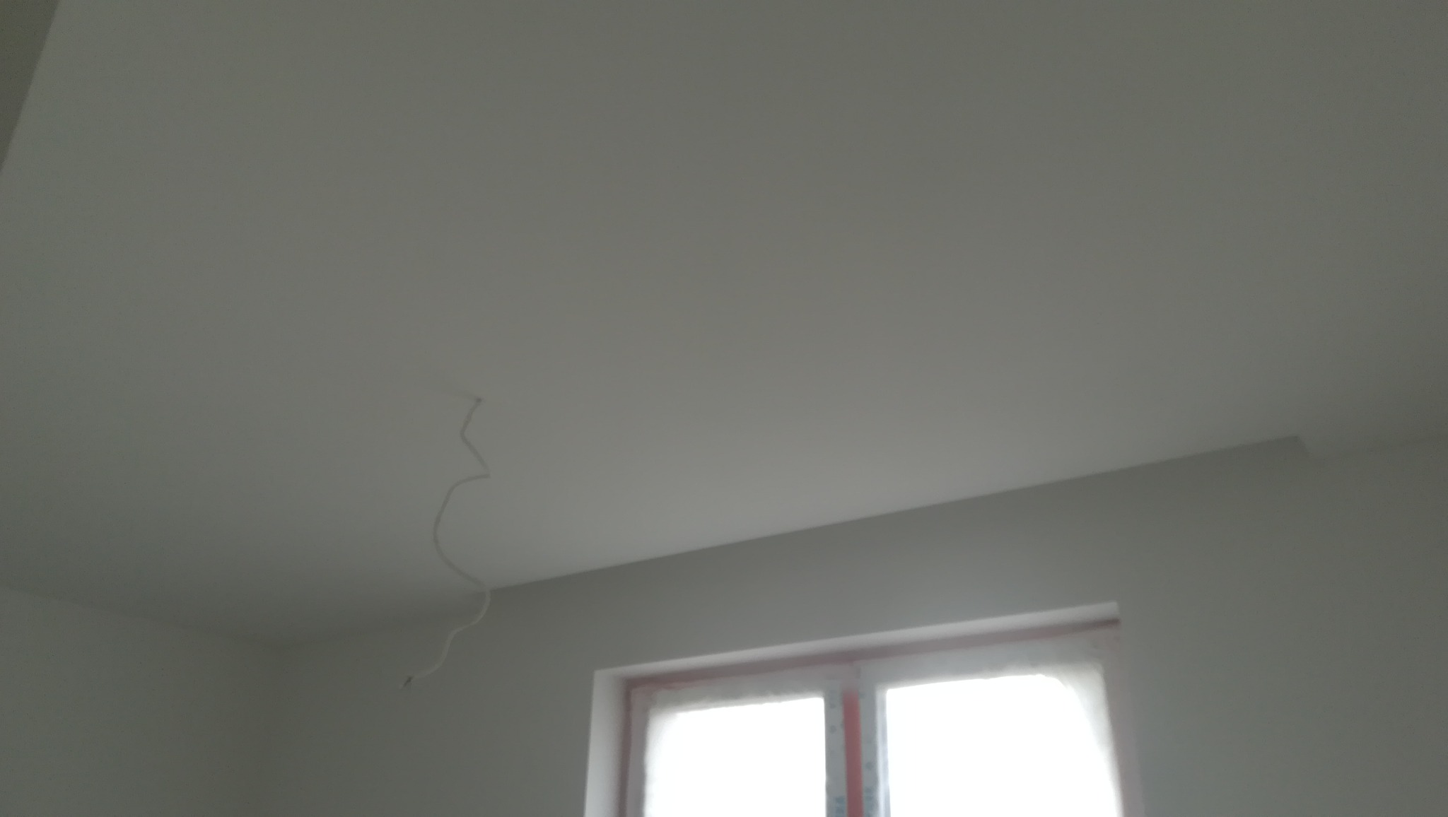 Gładka, biała powierzchnia sufitu z wystającym przewodem elektrycznym w rogu pokoju, widoczne okno w tle.
