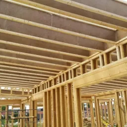 Wnętrze domu szkieletowego w trakcie budowy, widoczne drewniane belki konstrukcyjne i płyty OSB na suficie, z zewnątrz widoczne budynki w tle.