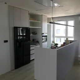 Nowoczesna kuchnia z białymi szafkami, czarną lod&oacute;wką side-by-side, wyspą kuchenną i minimalistycznym oświetleniem sufitowym.