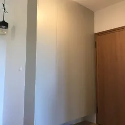 Wysoka, zabudowana szafa w przedpokoju, z parą białych sneakersów i ciemnych klapek u dołu, podświetlona wiszącą lampą z żarówką, obok drewniane drzwi.