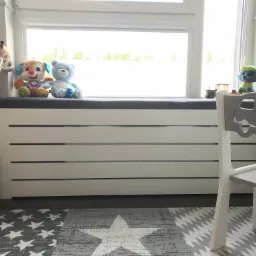 Biała zabudowa okienna z szarym siedziskiem w pokoju dziecięcym, ozdobiona pluszowymi zabawkami, obok dziecięce krzesełko z wyciętym kształtem samochodu i dywan w gwiazdy.