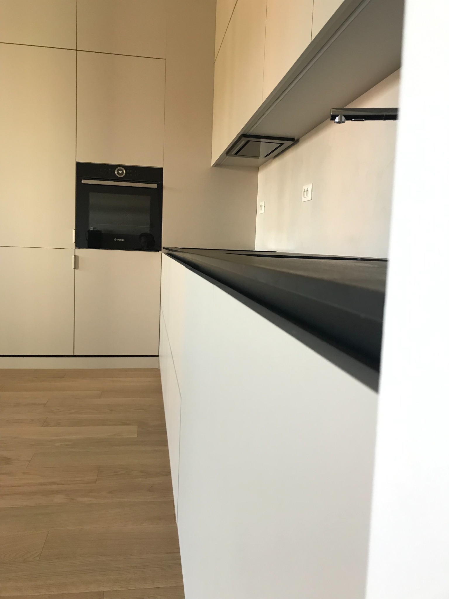 Minimalistyczna kuchnia z kremowymi szafkami, czarnym blatem i wbudowanym piekarnikiem Bosch, widoczna z perspektywy podłogi z jasnego drewna.