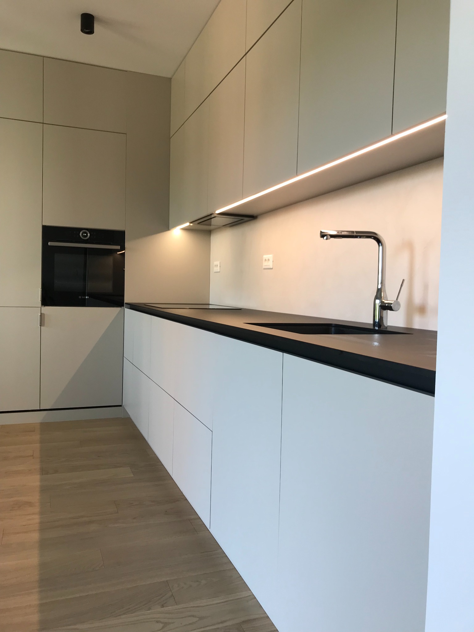 Minimalistyczna kuchnia z białymi szafkami dolnymi i szarymi górnymi, czarny blat, chromowana bateria, oświetlenie LED pod szafkami wiszącymi, drewniana podłoga.