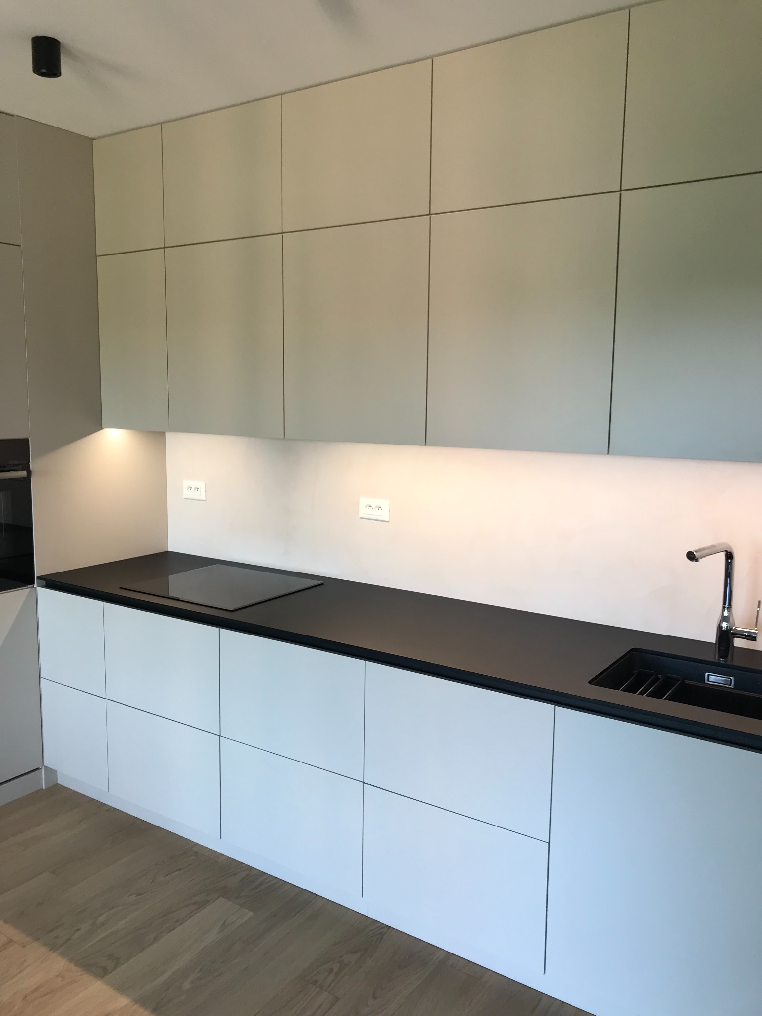 Minimalistyczna kuchnia z jasnymi, gładkimi frontami szafek, czarnym blatem i drewnianą podłogą, oświetlona punktowo pod górnymi szafkami.