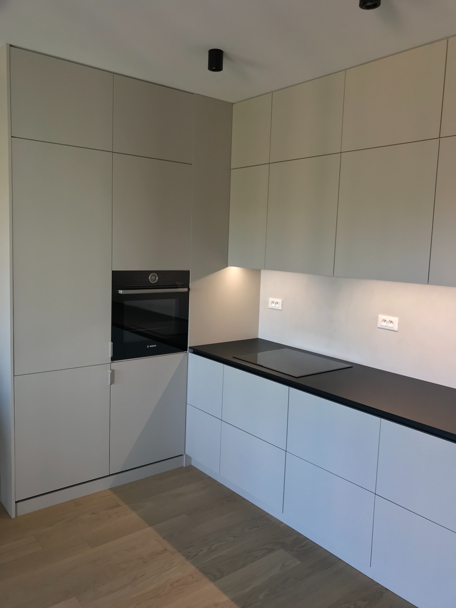 Minimalistyczna kuchnia z szarymi szafkami górnymi i białymi dolnymi, czarny blat, wbudowany piekarnik Bosch, płyta indukcyjna, drewniana podłoga, oświetlenie punktowe pod szafkami.