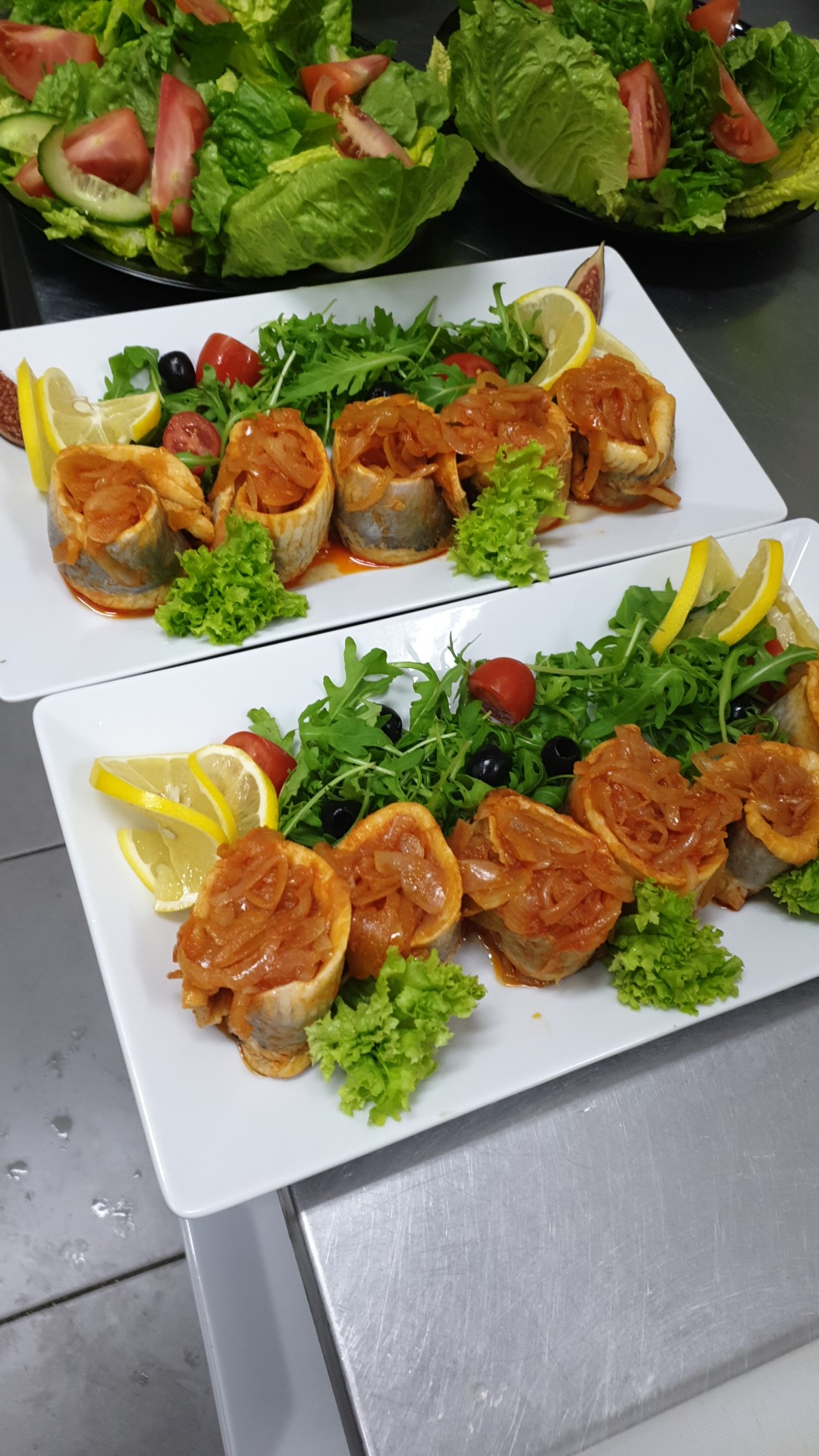 Dwie białe prostokątne tace z roladkami śledziowymi w sosie pomidorowo-cebulowym, udekorowane sałatą, rukolą, pomidorkami cherry, oliwkami i ćwiartkami cytryny, na tle sałatki warzywnej.