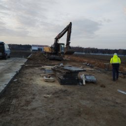 MAGOR-BUD Sp.z.o.o - Żółta koparka Volvo na placu budowy, obok sterta płyt betonowych, w tle budynek z napisem 'PŁYTY WARSTWOWE', pracownik w odblaskowej kamizelce idzie w stronę koparki.