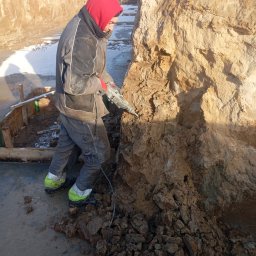 MAGOR-BUD Sp.z.o.o - Pracownik w czerwonej bluzie z kapturem, w okularach ochronnych, używa młota udarowego do obróbki ziemi w wykopie fundamentowym, obok widoczne drewniane szalunki i resztki śniegu.