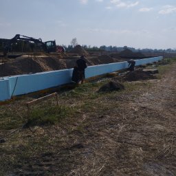 MAGOR-BUD Sp.z.o.o - Budowa fundamentów: pracownicy układają izolację termiczną z niebieskiego styropianu, widoczny wykop, ciężki sprzęt budowlany i hałdy piasku w tle.