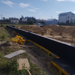 MAGOR-BUD Sp.z.o.o - Przygotowanie fundamentów: widok na wykop z zalanym betonem, zabezpieczony żółtymi panelami izolacyjnymi TASSU i czarną izolacją przeciwwilgociową, w tle hałdy piasku i zabudowa jednorodzinna...