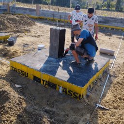 MAGOR-BUD Sp.z.o.o - Pracownik w rękawicach maluje pionowy element fundamentu czarną substancją, podstawa fundamentu zabezpieczona żółtą folią TASSU, w tle dwaj obserwujący pracownicy i teren budowy.