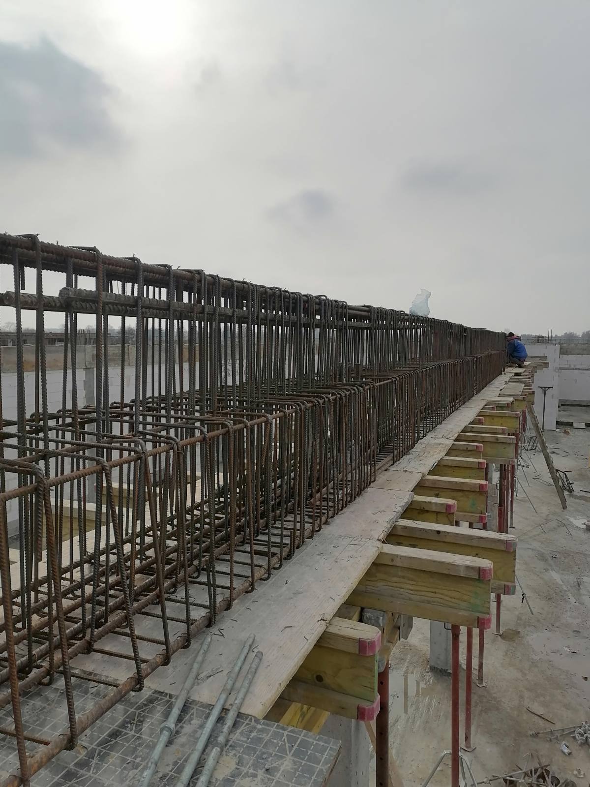 Zbrojenie pod wylewkę stropu na placu budowy, widoczne drewniane podpory szalunku i pracownik w oddali.