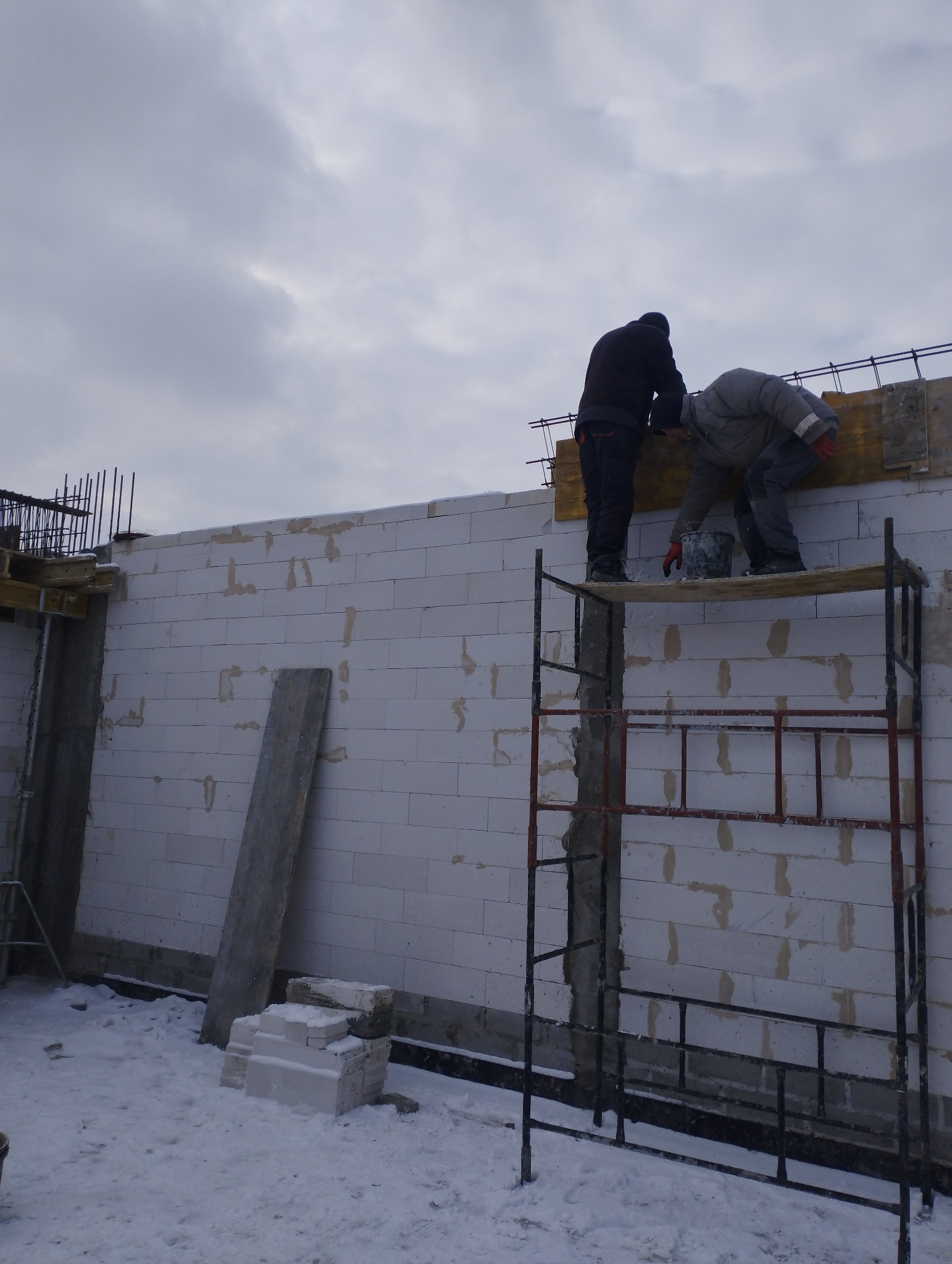 Dwóch murarzy na rusztowaniu, pracujących nad wznoszeniem ściany z białych bloczków w zimowych warunkach, widoczne zbrojenie i częściowe wypełnienie spoin.