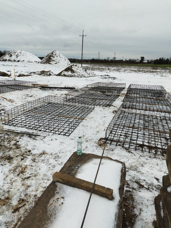 Zbrojenie fundamentów w kształcie prostokątnych klatek ułożone na śniegu, w tle pryzmy ziemi pod śniegiem i słup energetyczny, butelka wody na betonowym elemencie w pierwszym planie.
