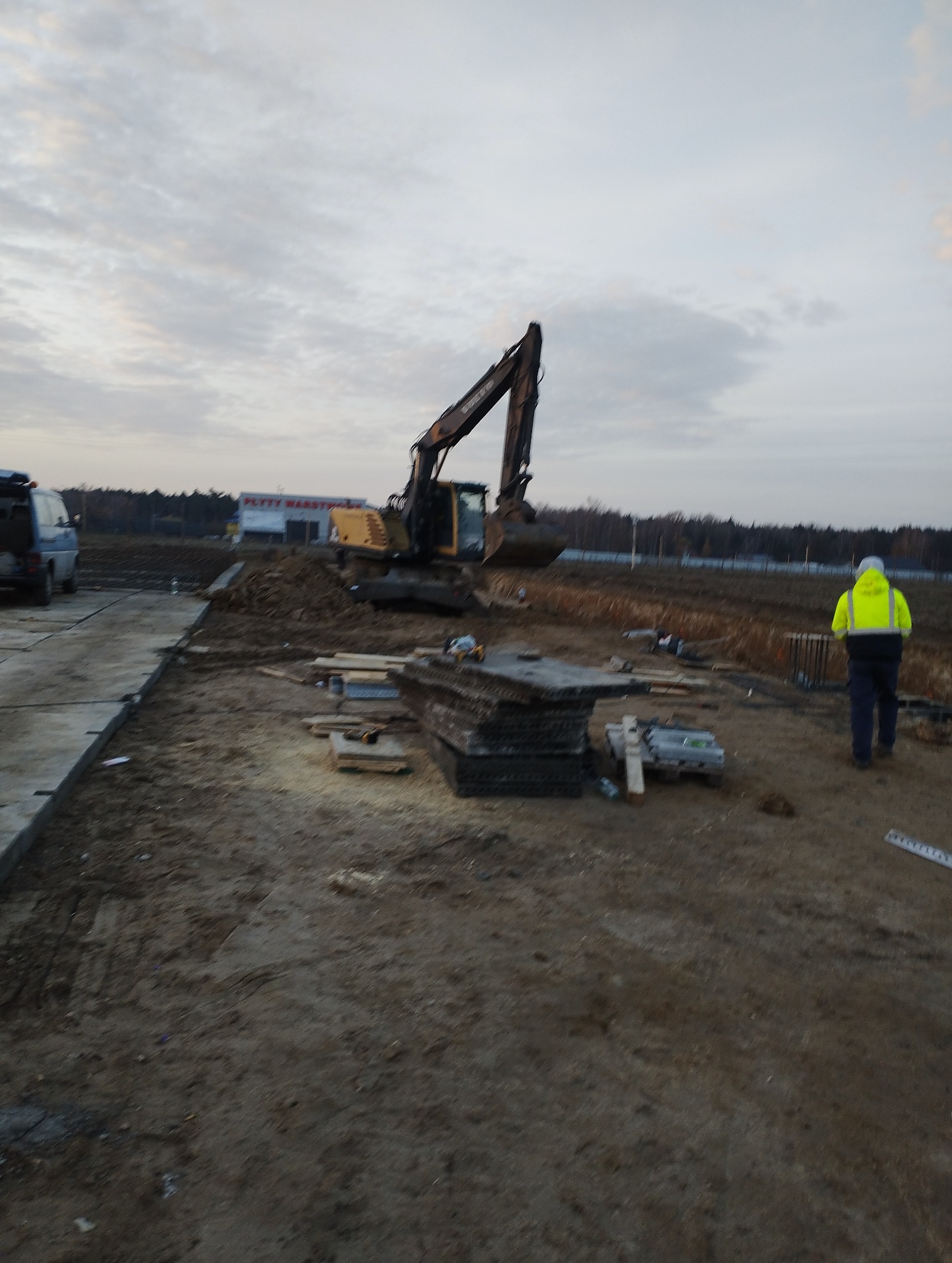 Żółta koparka Volvo na placu budowy, obok sterta płyt betonowych, w tle budynek z napisem 'PŁYTY WARSTWOWE', pracownik w odblaskowej kamizelce idzie w stronę koparki.