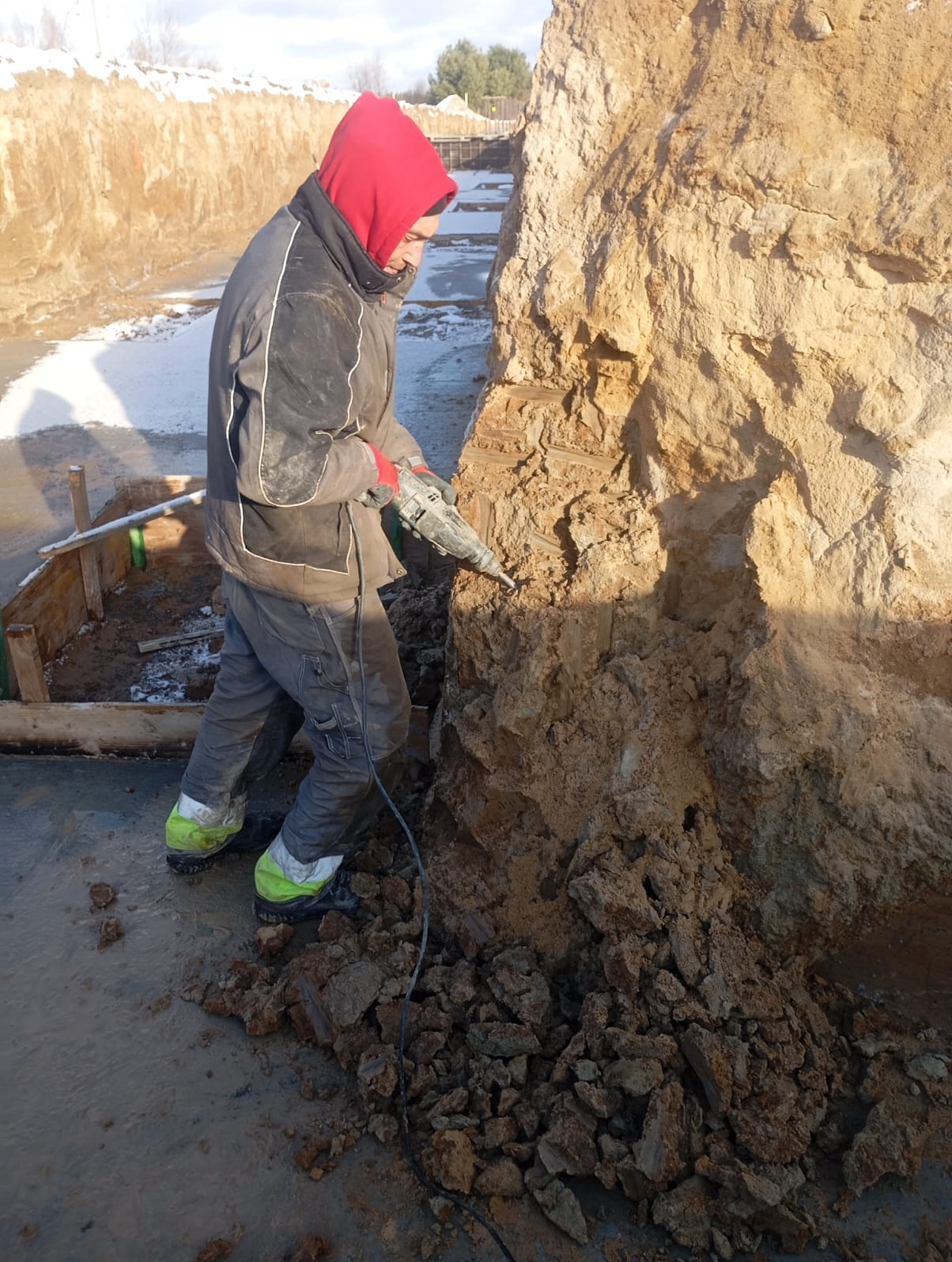 Pracownik w czerwonej bluzie z kapturem, w okularach ochronnych, używa młota udarowego do obróbki ziemi w wykopie fundamentowym, obok widoczne drewniane szalunki i resztki śniegu.