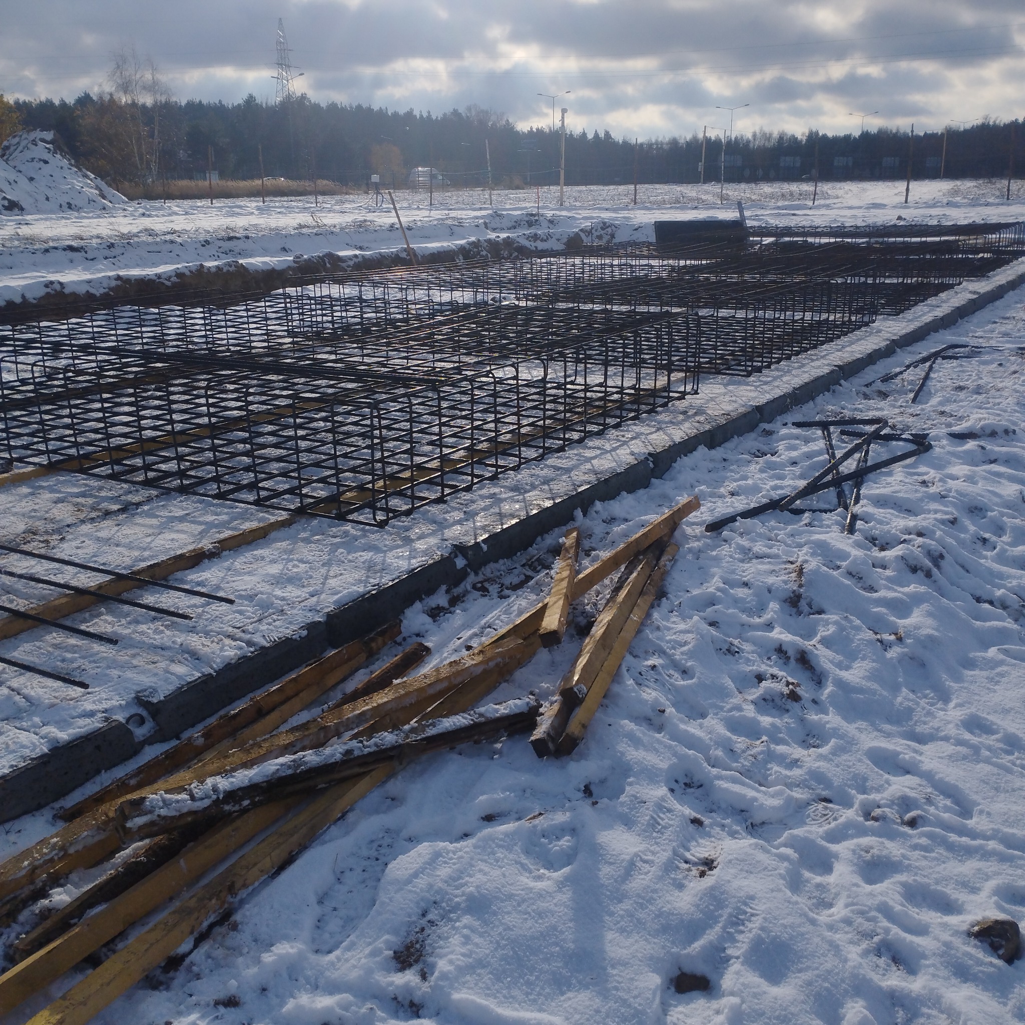 Zbrojenie fundamentów w zasypanym śniegiem wykopie, widoczne drewniane podpory i fragmenty szalunków, w tle drzewa i słupy energetyczne.