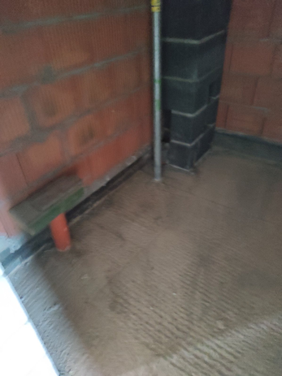 Surowa, świeżo wylana podłoga w pomieszczeniu z ceglanymi ścianami i fragmentem komina, widoczny pionowy element instalacji i fragment deski oparty o ścianę.