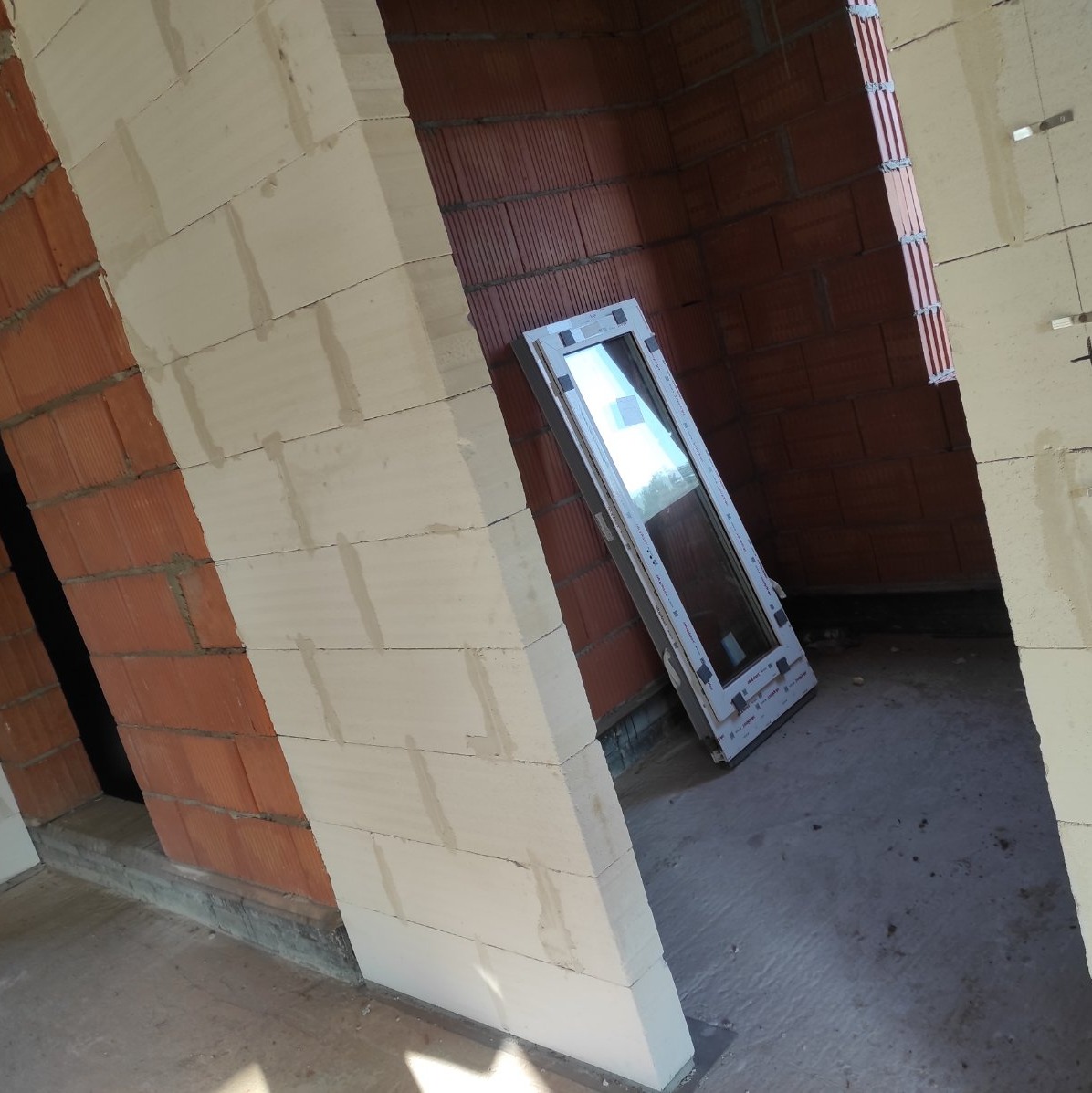 Wnętrze domu w trakcie budowy: ściany z pustaków ceramicznych, okno PCV oparte o ścianę, surowy beton na podłodze, widoczne fragmenty instalacji elektrycznej.