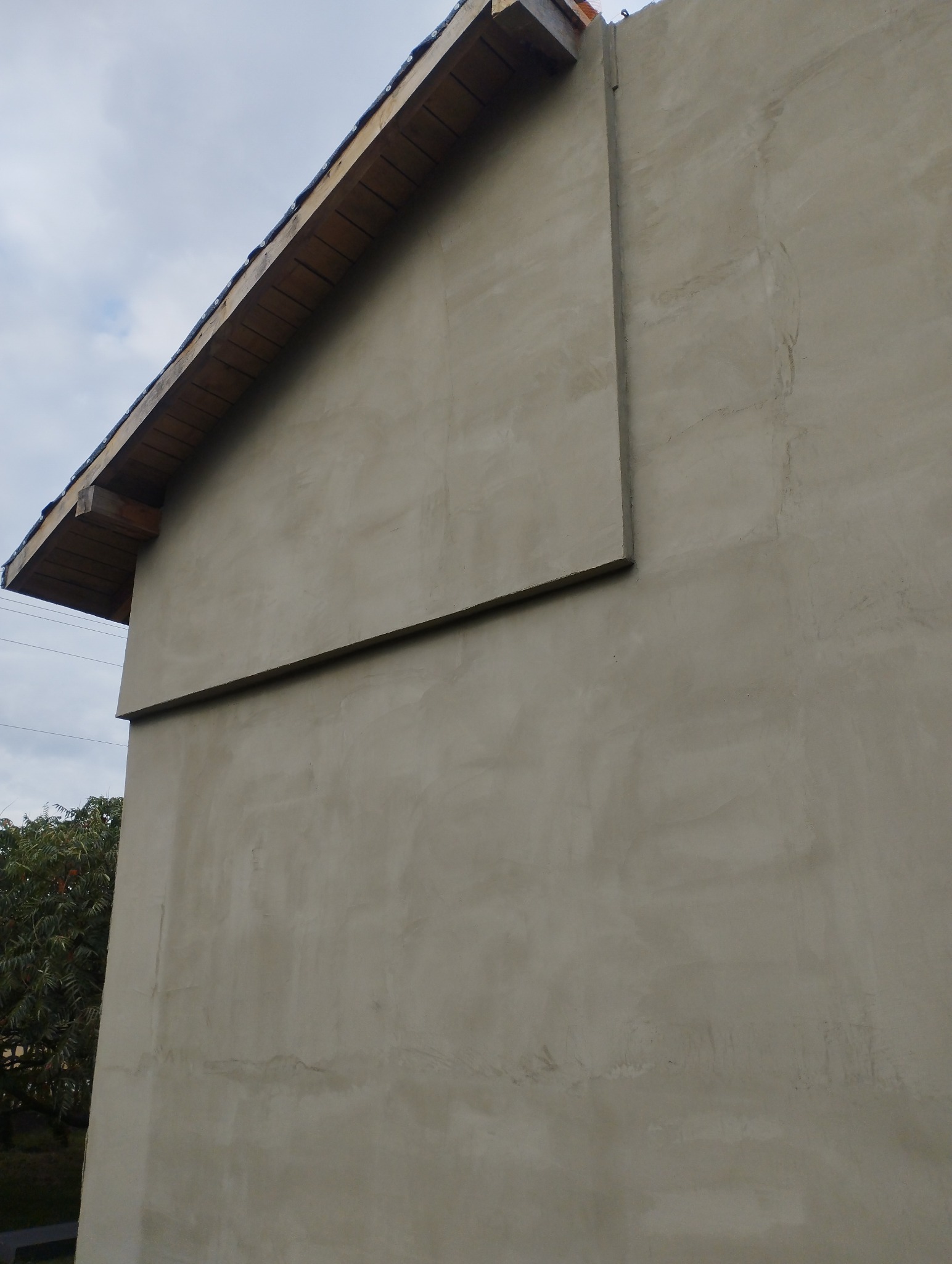 Świeżo otynkowana ściana domu z widoczną konstrukcją dachu z drewnianymi belkami i fragmentem elewacji z nierówną, szarą fakturą tynku. Widoczny fragment drzewa w tle.