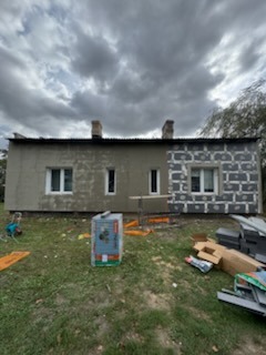 Budynek jednokondygnacyjny w trakcie ocieplania. Fragment ściany z widoczną warstwą izolacji termicznej w postaci szarych płyt, pozostała część ściany pokryta szarym tynkiem. Na pierwszym planie...