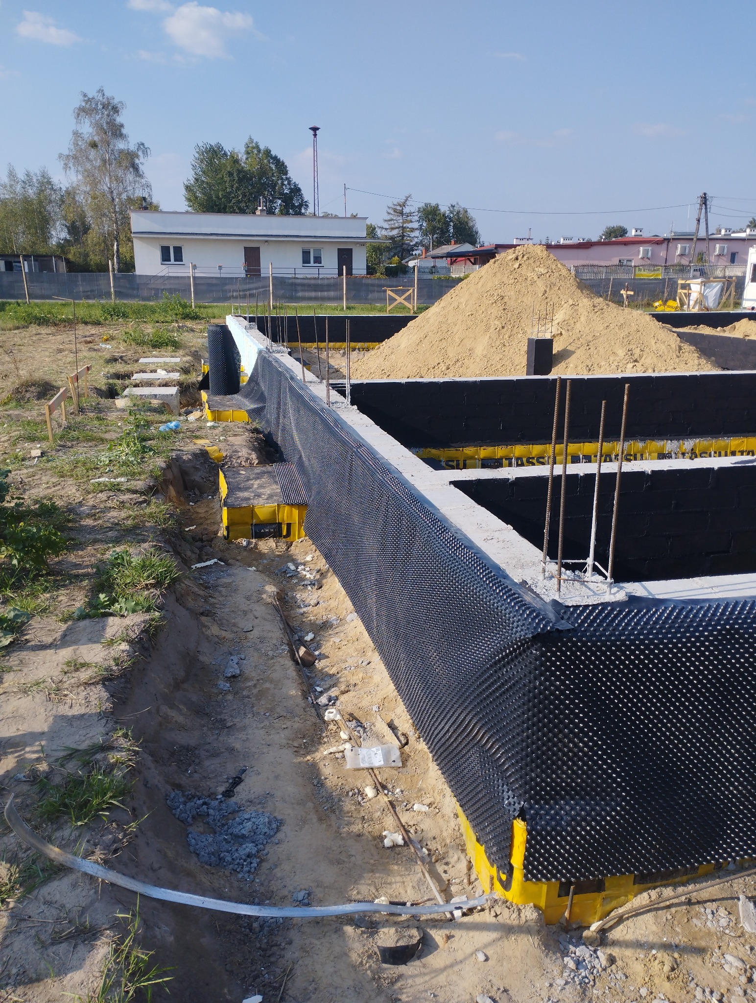 Fundamenty domu w trakcie budowy, zabezpieczone folią kubełkową i żółtymi elementami szalunkowymi, z widocznym wykopem i stertą piasku w tle.