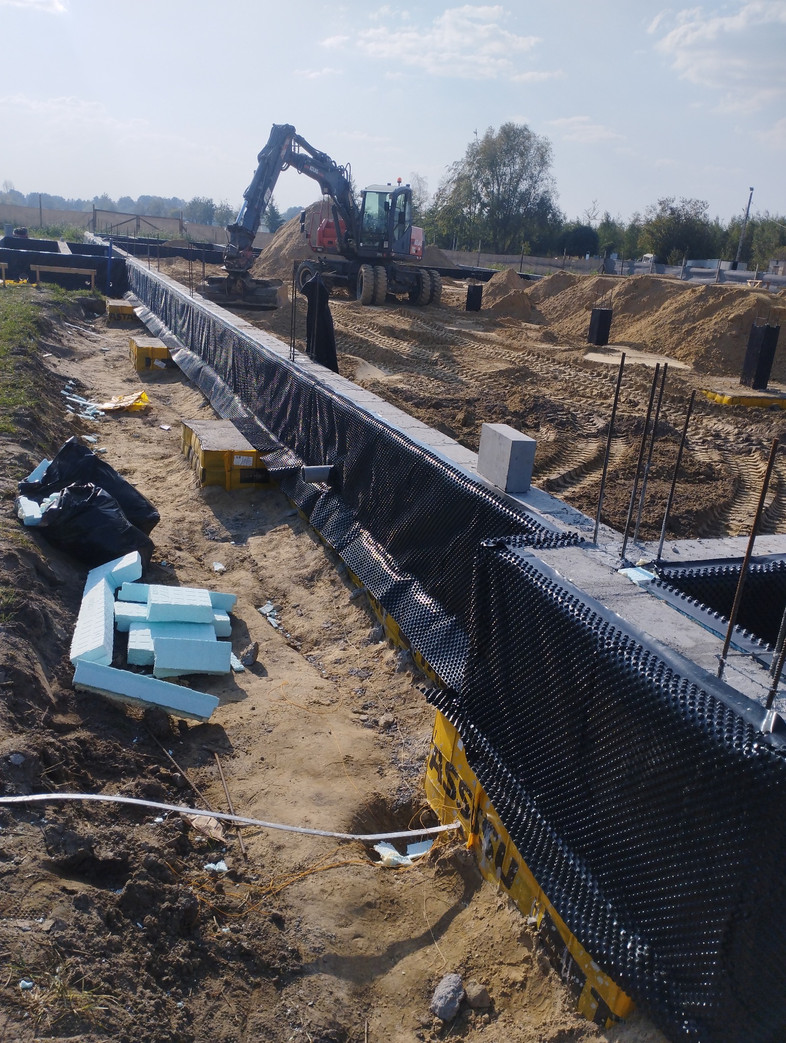 Fundament z izolacją pionową z folii kubełkowej i przygotowanym ociepleniem ze styropianu, w tle koparka na placu budowy.