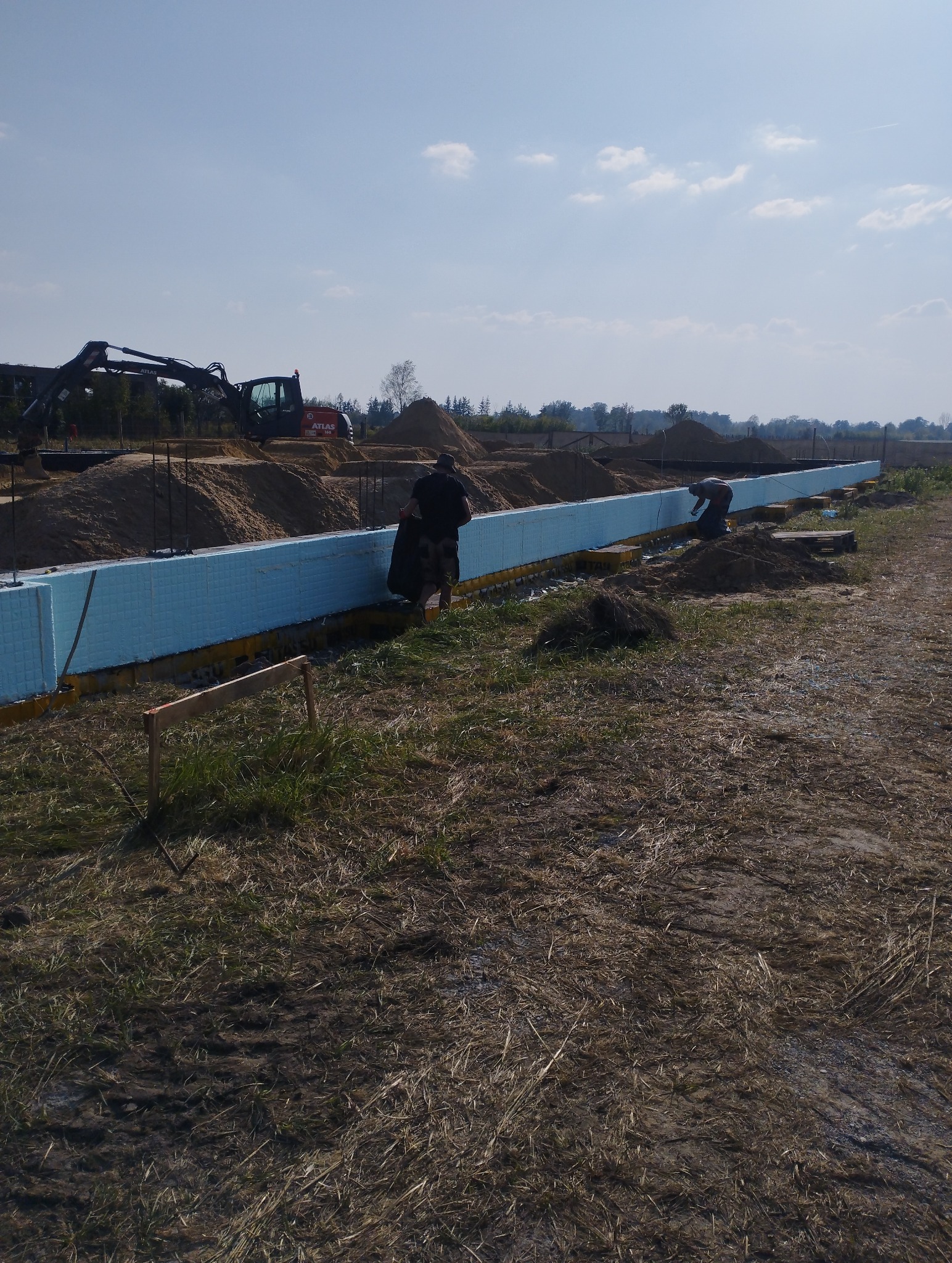 Budowa fundamentów: pracownicy układają izolację termiczną z niebieskiego styropianu, widoczny wykop, ciężki sprzęt budowlany i hałdy piasku w tle.