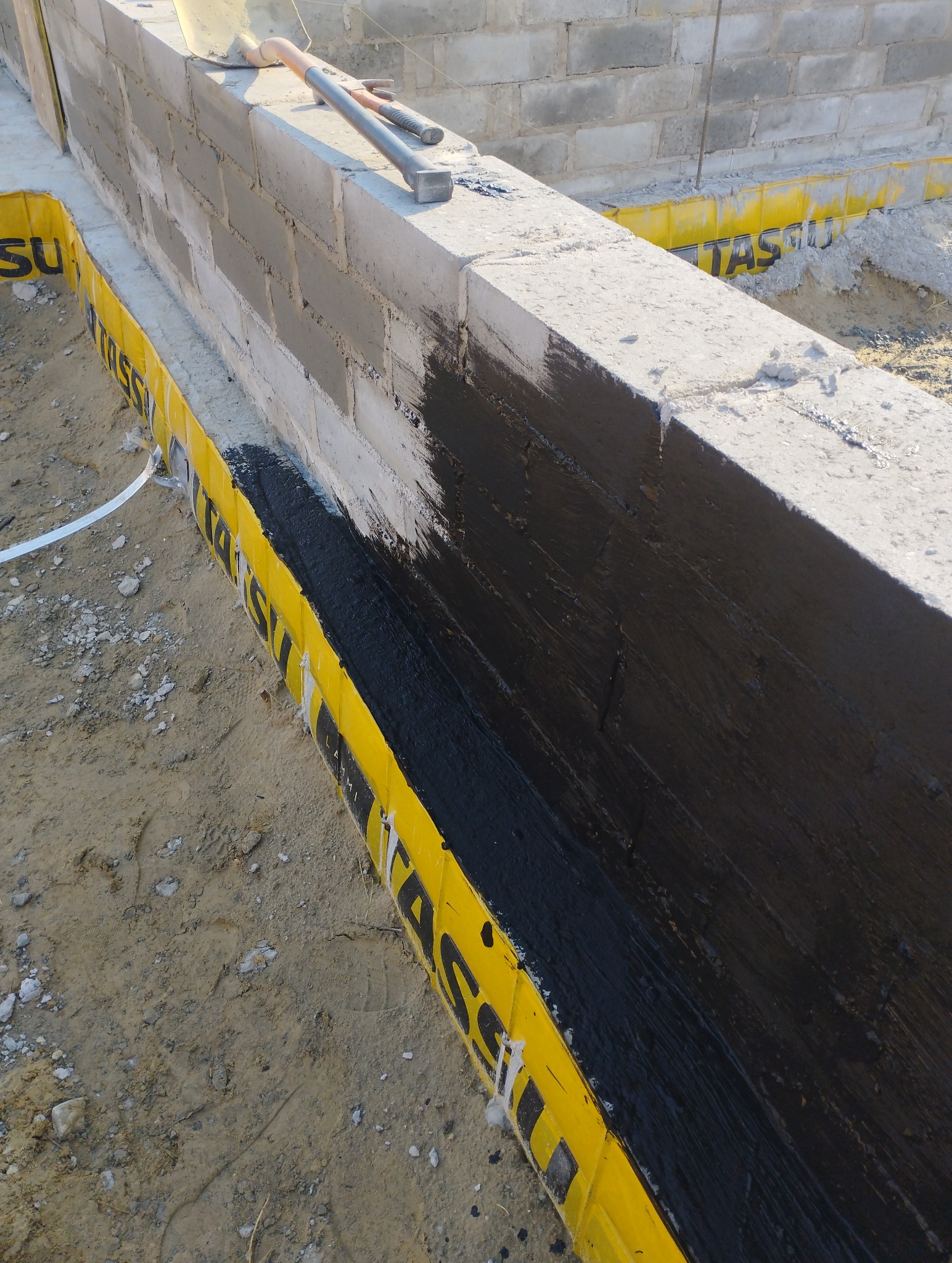 Świeżo zaizolowany fundament z bloczków betonowych, zabezpieczony czarną masą bitumiczną i żółtą folią ochronną, na wierzchu leżą narzędzia murarskie.