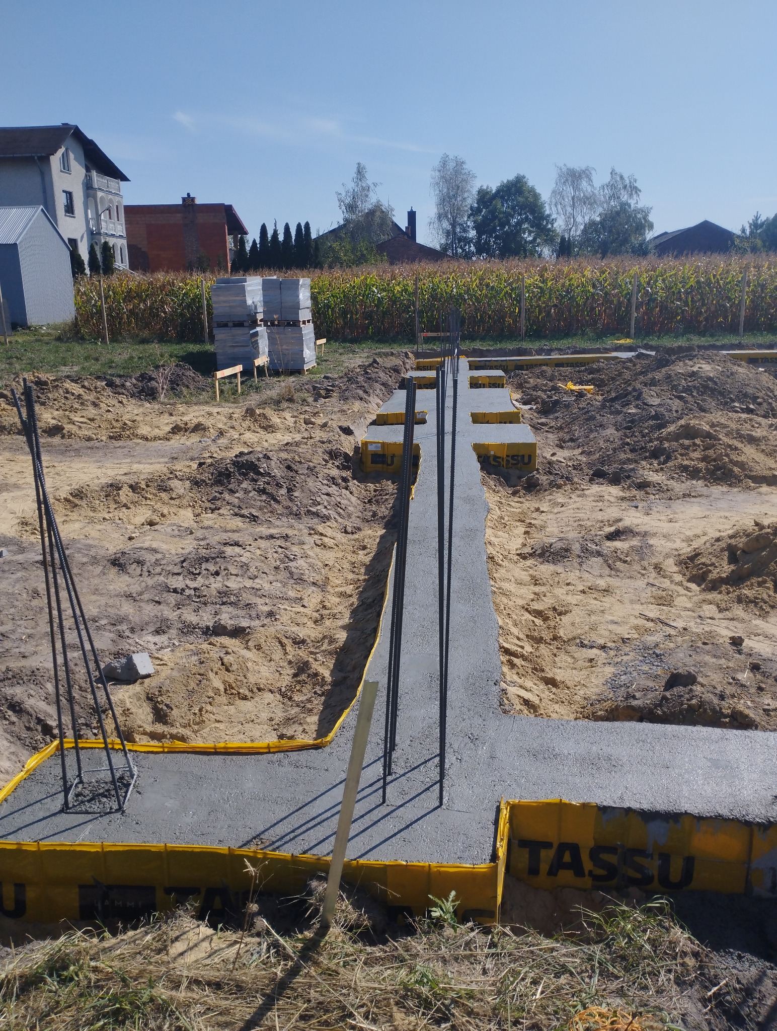 Wylany fundament w kształcie litery L z wystającymi prętami zbrojeniowymi, zabezpieczony żółtą izolacją przeciwwilgociową TASSU, na tle pola kukurydzy i zabudowy jednorodzinnej w słoneczny dzień.