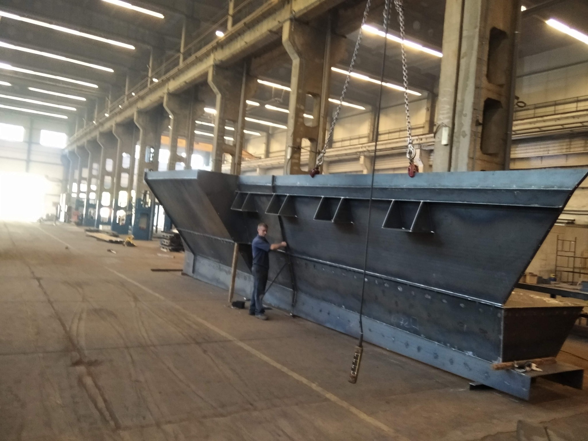 Duża, surowa, stalowa konstrukcja z licznymi spoinami, zawieszona na łańcuchach w hali produkcyjnej. Pracownik w niebieskiej koszuli stoi obok, wykonując prace wykończeniowe.