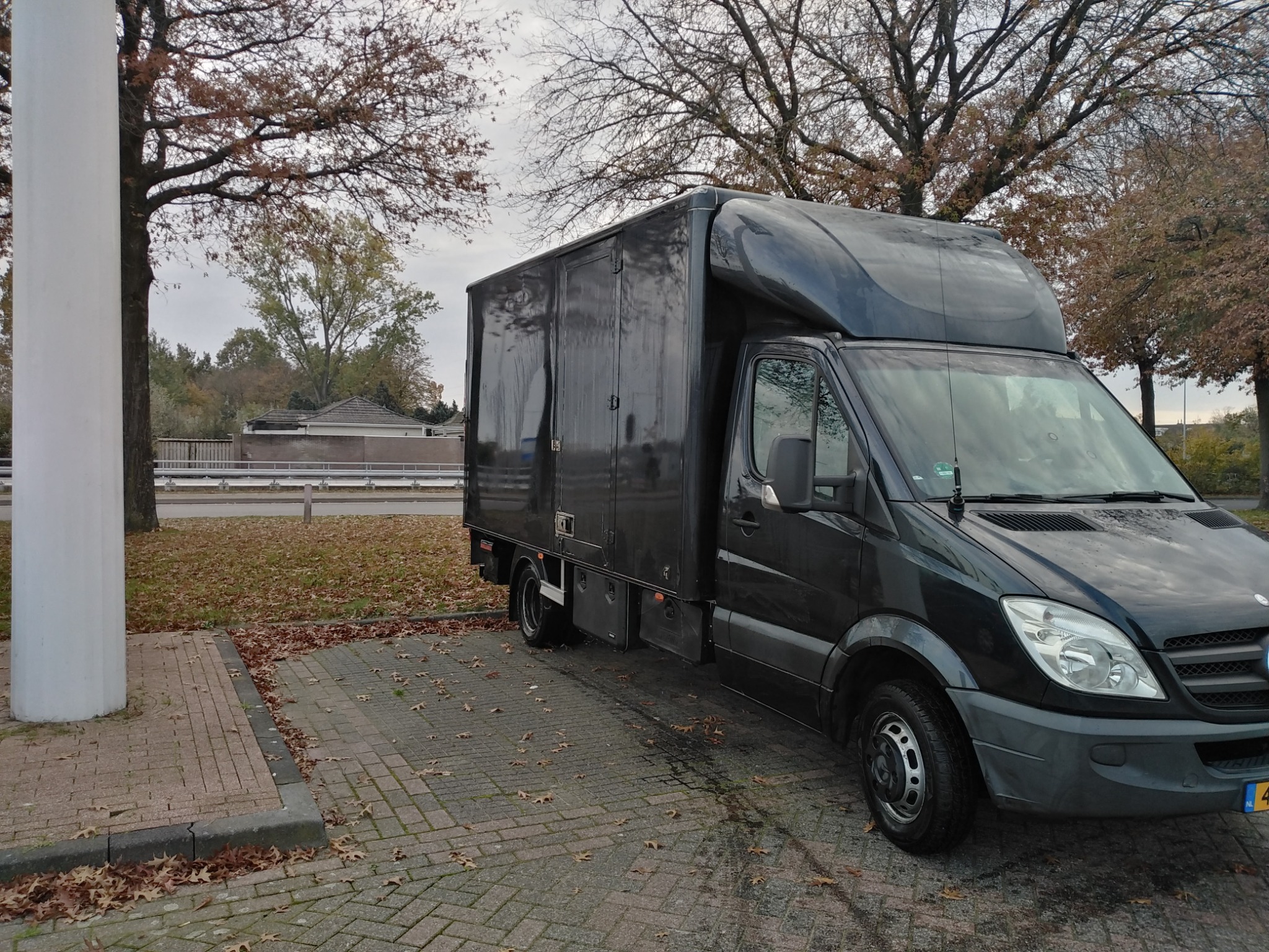 Czarny bus dostawczy Mercedes-Benz Sprinter z wysoką zabudową, zaparkowany na brukowanym parkingu w jesiennej scenerii z opadłymi liśćmi.