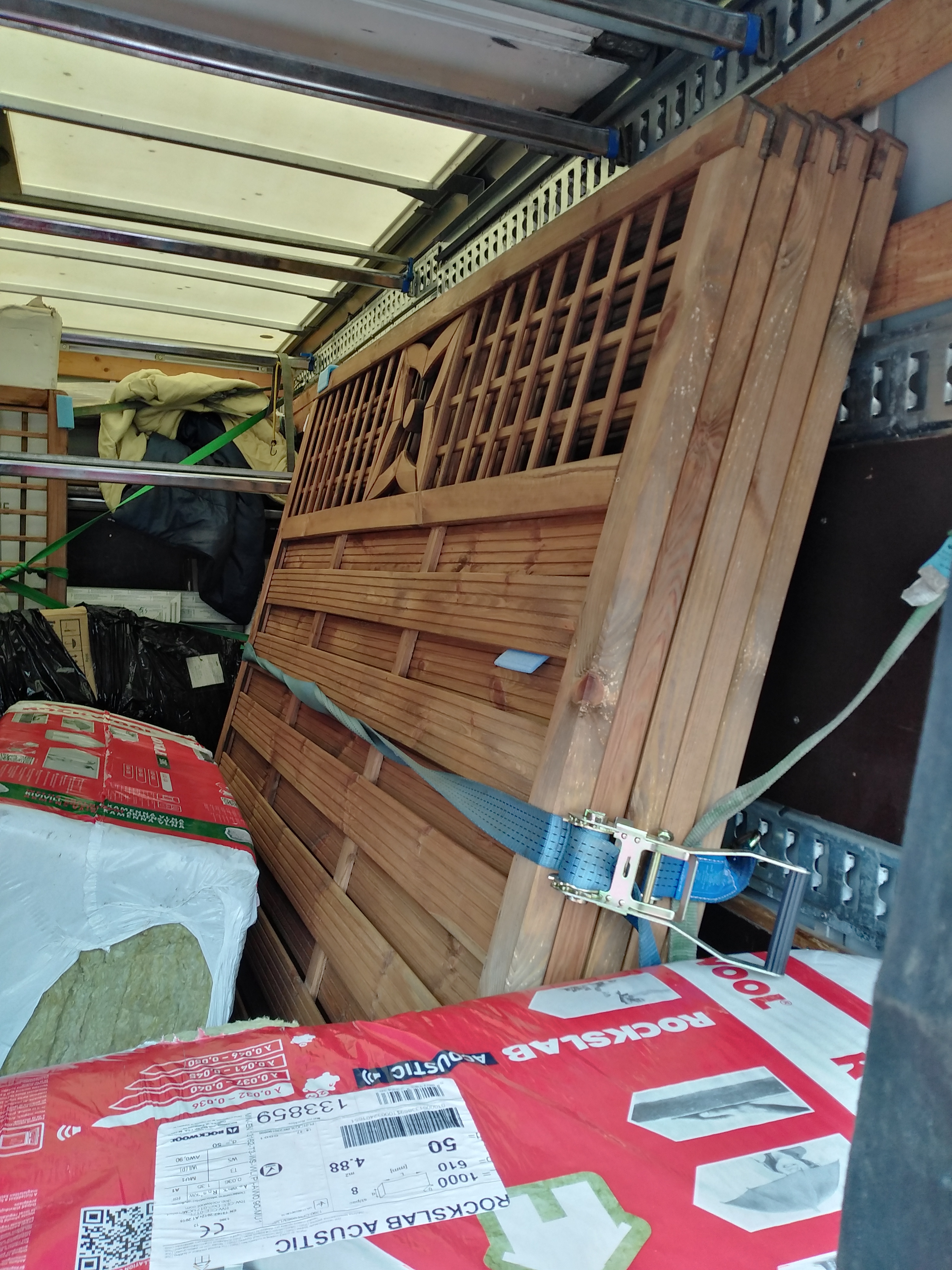 Drewniane panele ogrodzeniowe zabezpieczone pasem transportowym wewnątrz pojazdu dostawczego, obok paczki wełny mineralnej Rockslab Acustic.