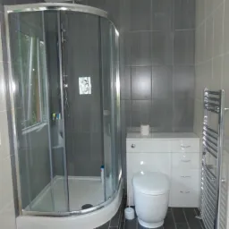 Nowoczesna łazienka z kabiną prysznicową z zaokrąglonym szkłem, szare płytki ścienne i podłogowe, biała toaleta z szafką i grzejnik drabinkowy.