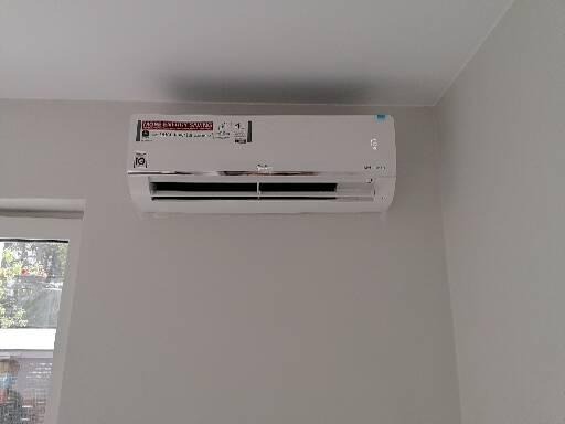 Biała klimatyzacja ścienna LG zamontowana w jasnym pomieszczeniu z widokiem na zieleń przez okno.