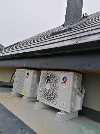 Dwa białe agregaty klimatyzacji marki Gree zamontowane na dachu budynku z szarą dachówką, widoczne przewody.