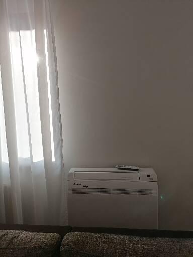 Biała klimatyzacja przenośna marki Kaisai stoi przy ścianie obok okna z białymi zasłonami i pilotem na górze.