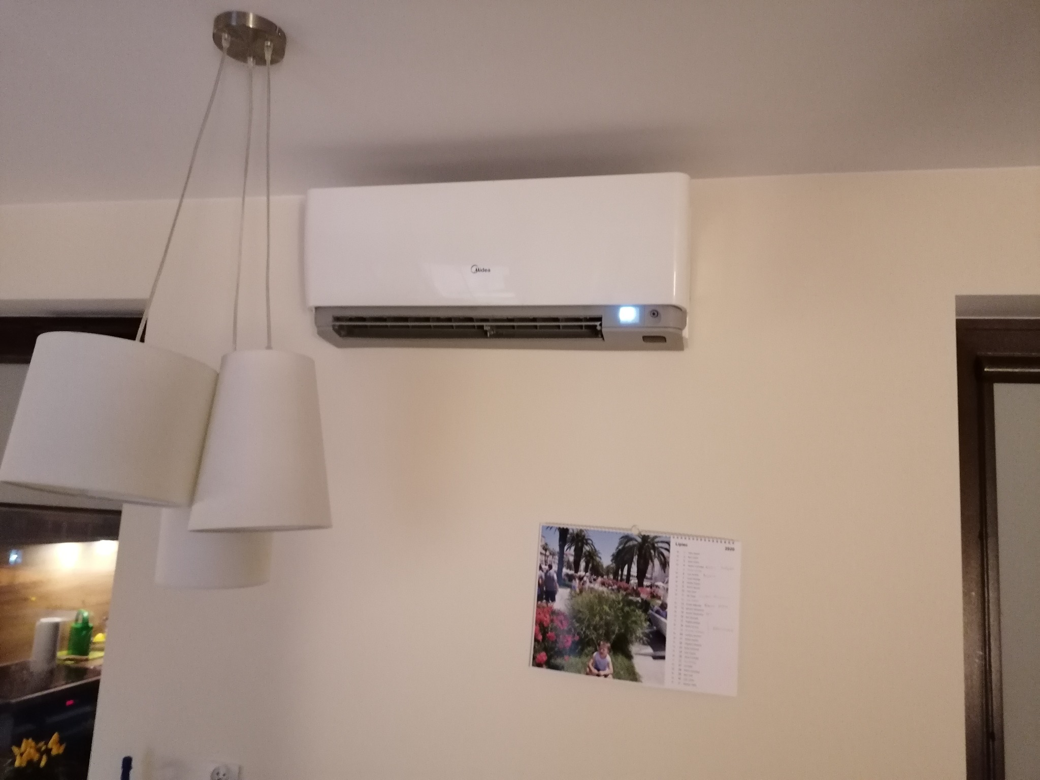 Biały klimatyzator ścienny marki Midea zamontowany w jasnym pokoju, widoczny fragment lampy wiszącej i kalendarz na ścianie.