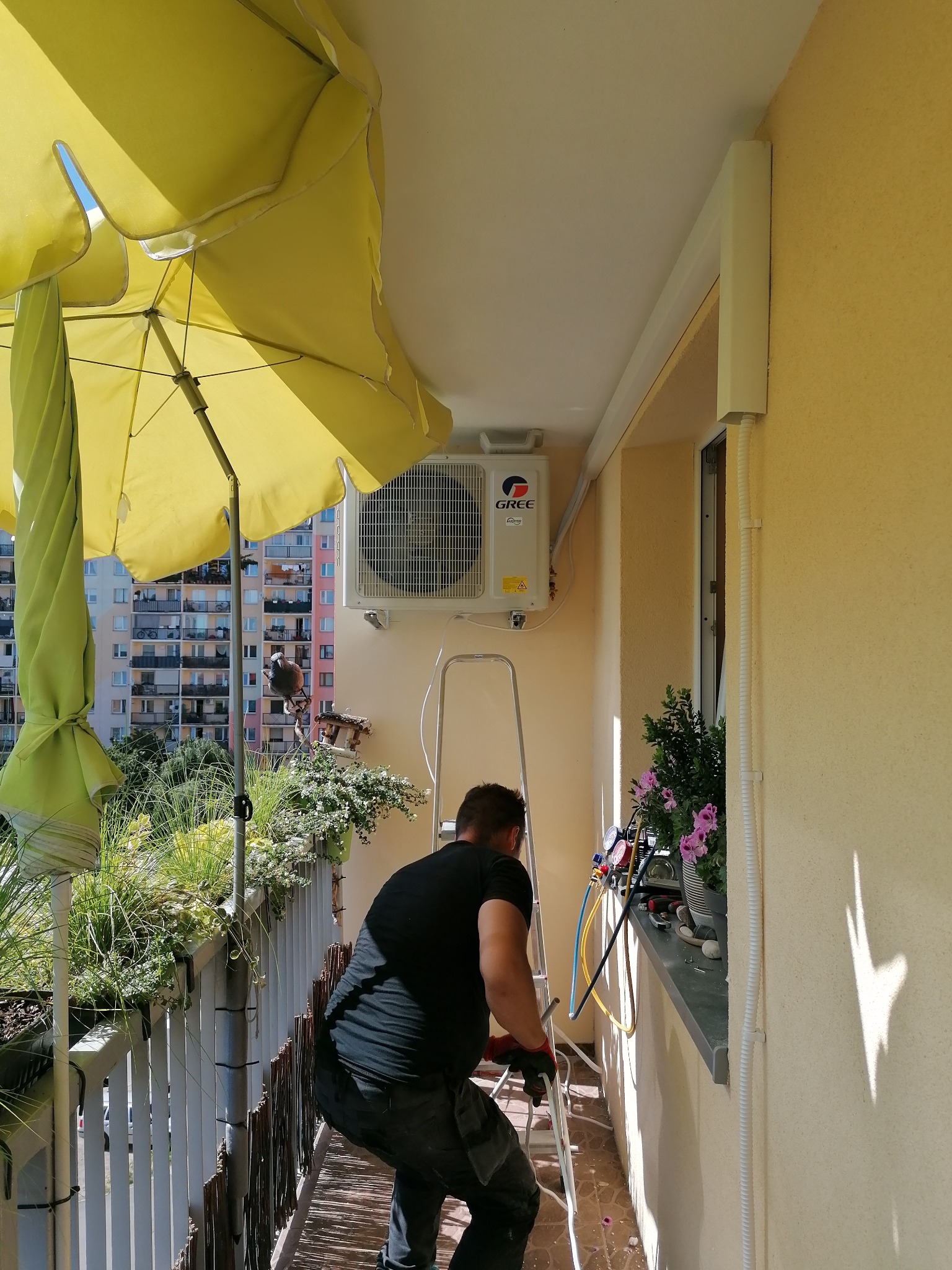 Instalator na balkonie, na drabinie, pod żółtym parasolem, montuje klimatyzator marki Gree na ścianie budynku wielorodzinnego, widoczne narzędzia i manometr.