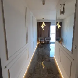 Nowoczesny korytarz z ciemną, marmurkową posadzką, oświetleniem LED wzdłuż ścian z dekoracyjnymi panelami i lustrem, zakończony pokojem z dwoma szarymi fotelami.