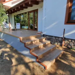 Zewnętrzne schody i taras wyłożone płytkami imitującymi drewno, z brązowymi listwami wykończeniowymi, widok z dołu na dom z białymi ścianami i brązowymi oknami, obok drzewo owocowe i teren...