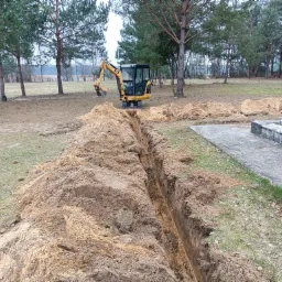 Wąski wykop w ziemi, z żółtą mini koparką CAT w tle, otoczony stertami ziemi i drzewami.