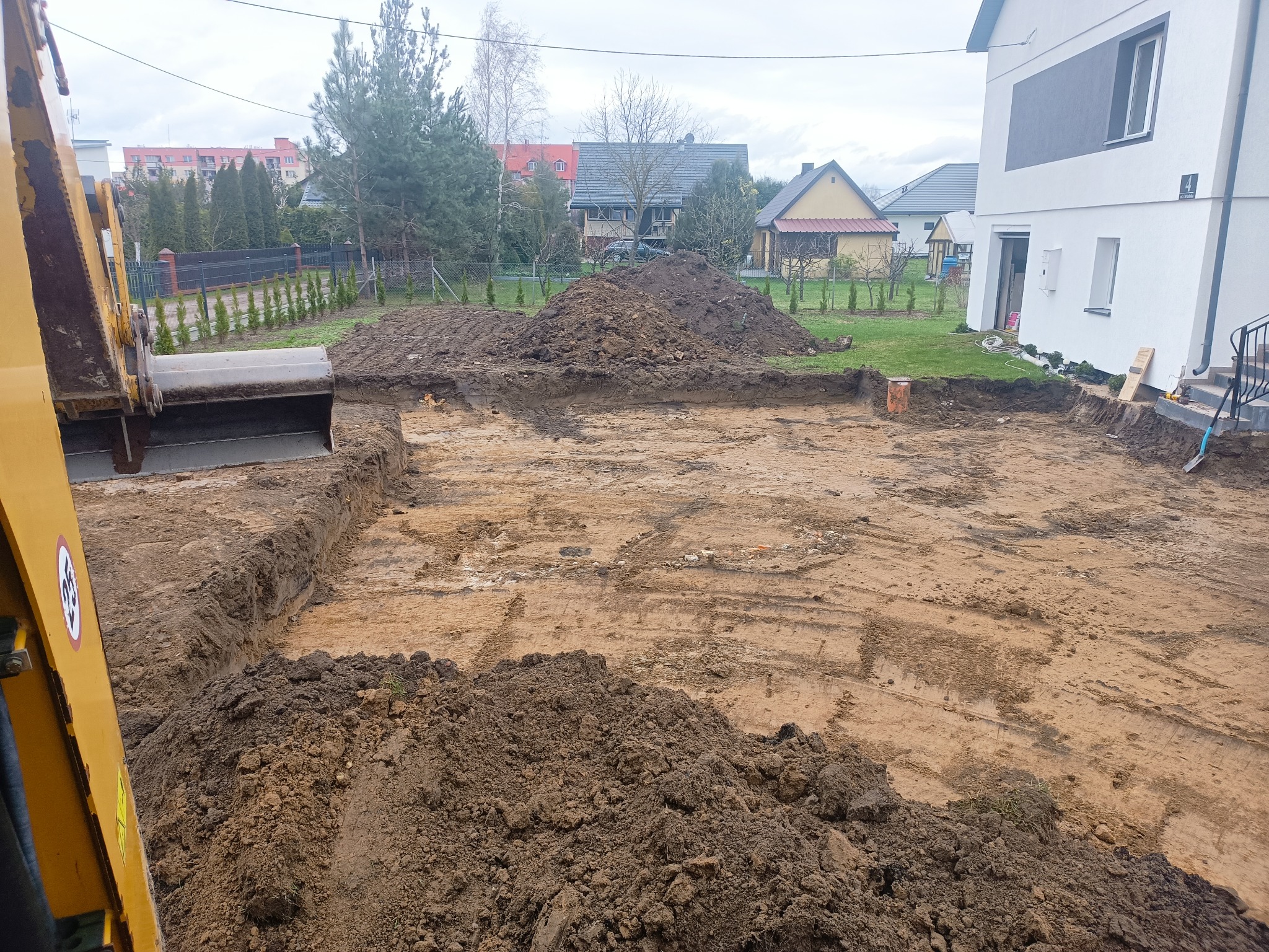 Żółta koparka przy wykopie pod fundamenty w Bielsku Podlaskim, widoczne ułożone warstwy ziemi i hałda wykopanej ziemi, w tle dom w zabudowie jednorodzinnej.