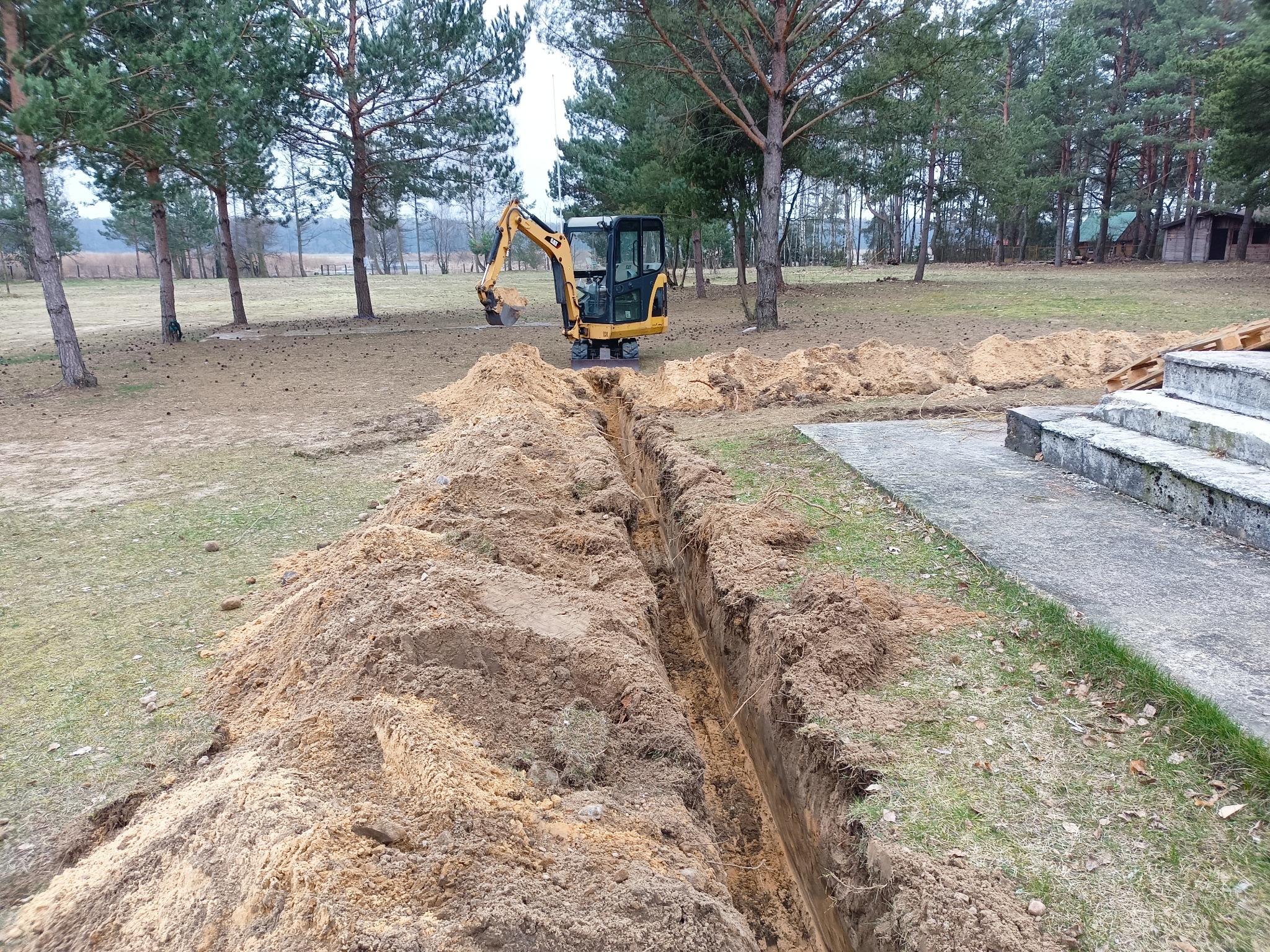 Wąski wykop w ziemi, z żółtą mini koparką CAT w tle, otoczony stertami ziemi i drzewami.