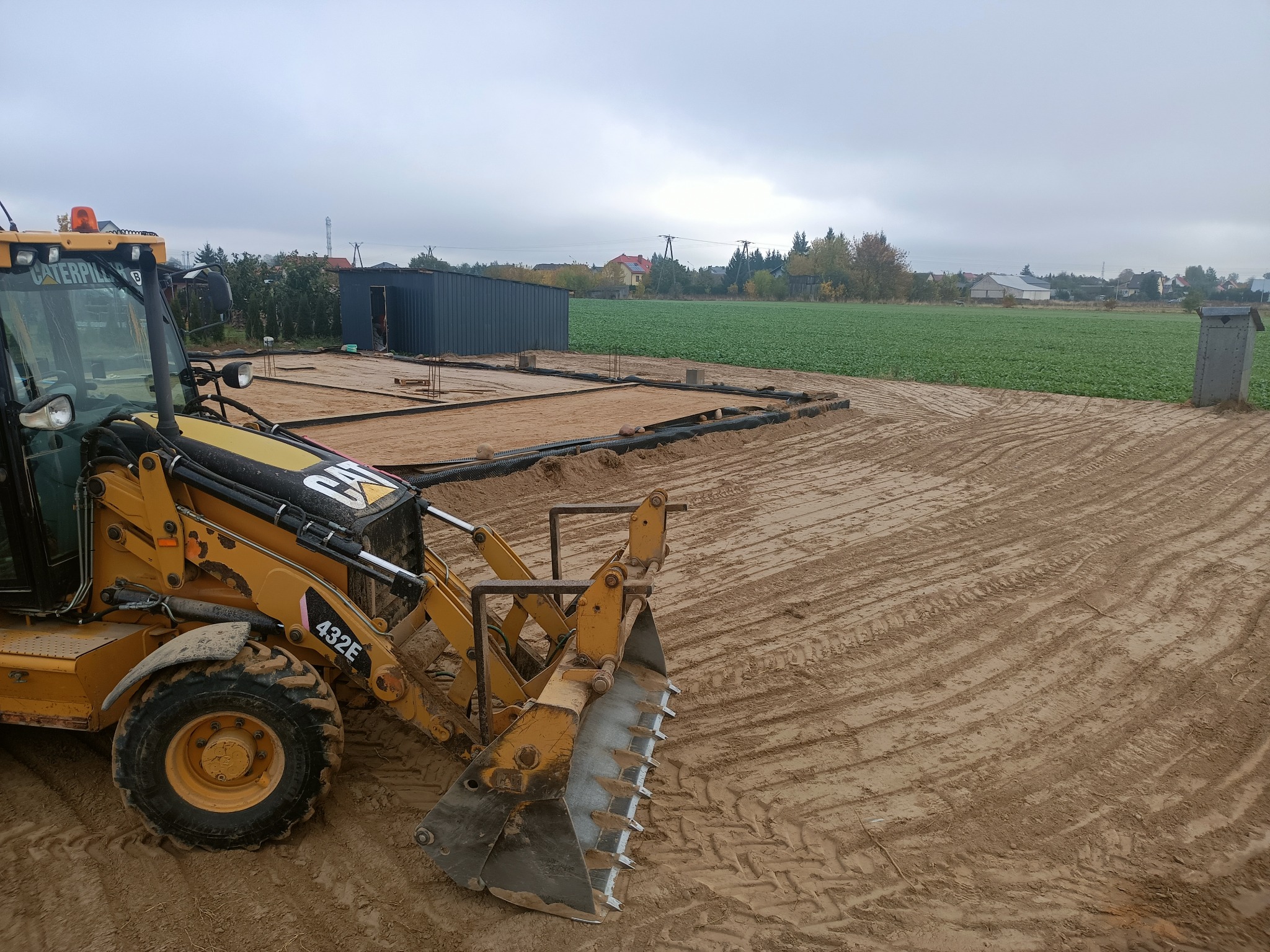 Żółta koparko-ładowarka Caterpillar 432E na piaszczystym terenie z przygotowanymi fundamentami, widoczny szary magazyn blaszany i pole uprawne w tle, pochmurne niebo.